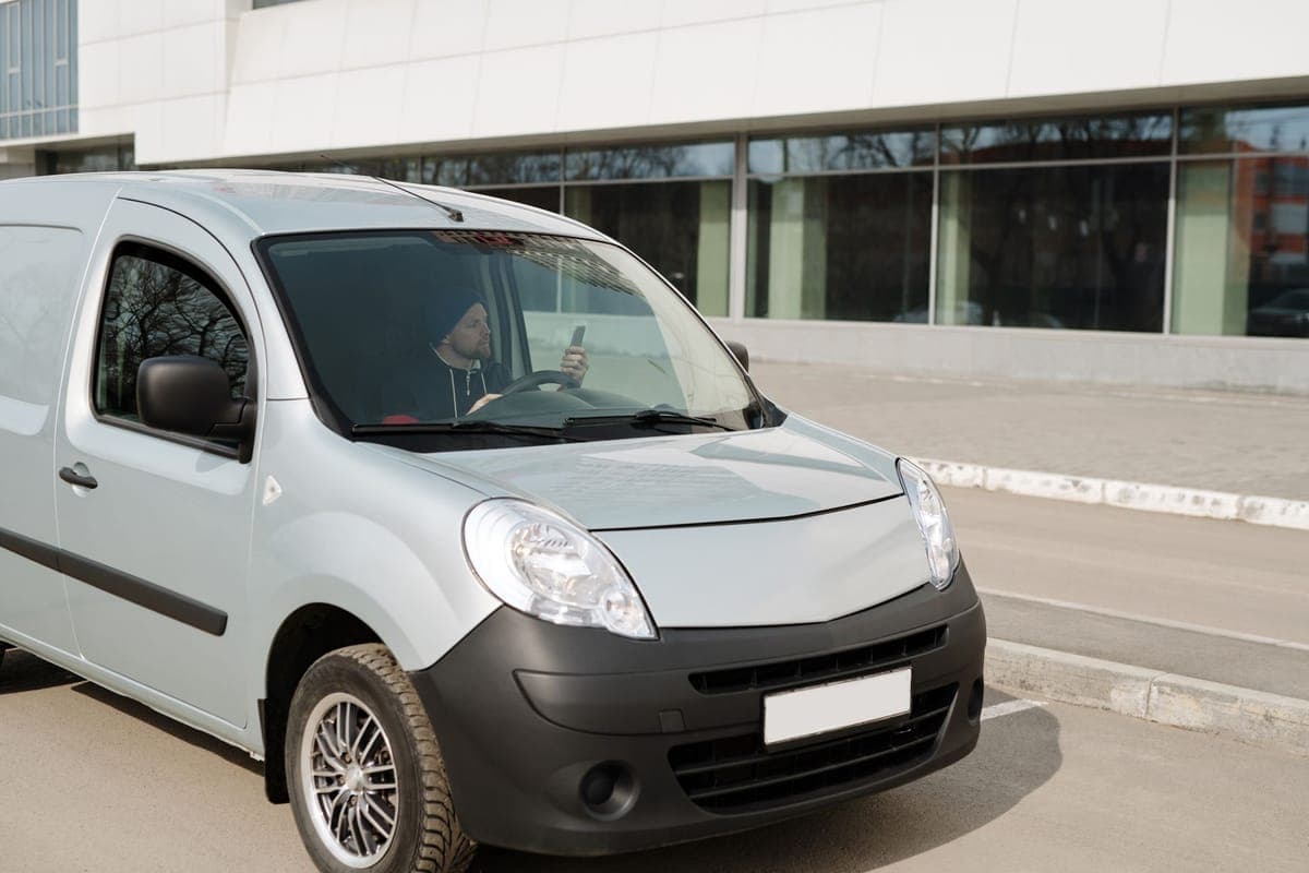 A driver holding a phone at the wheel of a small white van parked outside a building