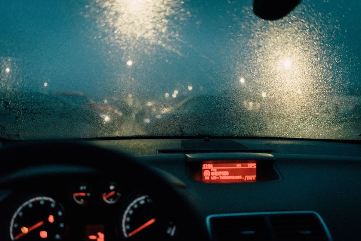 A rain-covered windshield at night, with oncoming headlights blown into bright blobs above a dimly lit dashboard