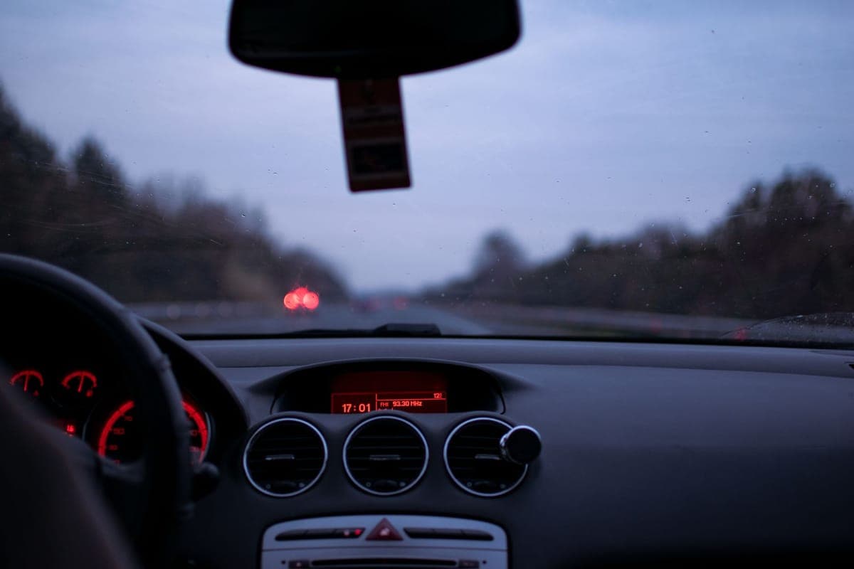 A car windshield and lit instrument panel at dusk, with the tail lights of traffic ahead